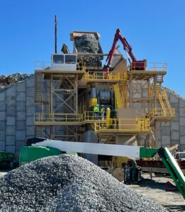 A large industrial facility processes a massive pile of aggregate. Construction equipment and workers are visible around the structure, while the control cab monitors activities under a clear blue sky.