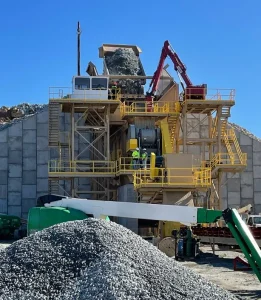 A large industrial facility processes a massive pile of aggregate. Construction equipment and workers are visible around the structure, while the control cab monitors activities under a clear blue sky.